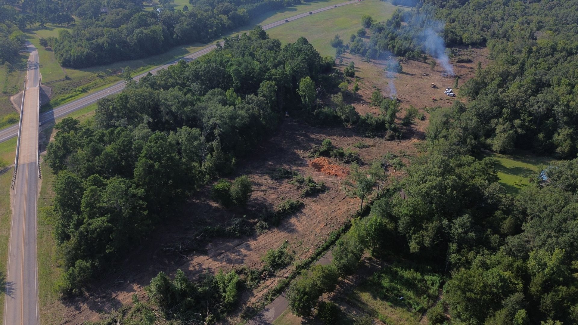 Aerial view: Trees surround a clearing with smoke, near a road and bridge.