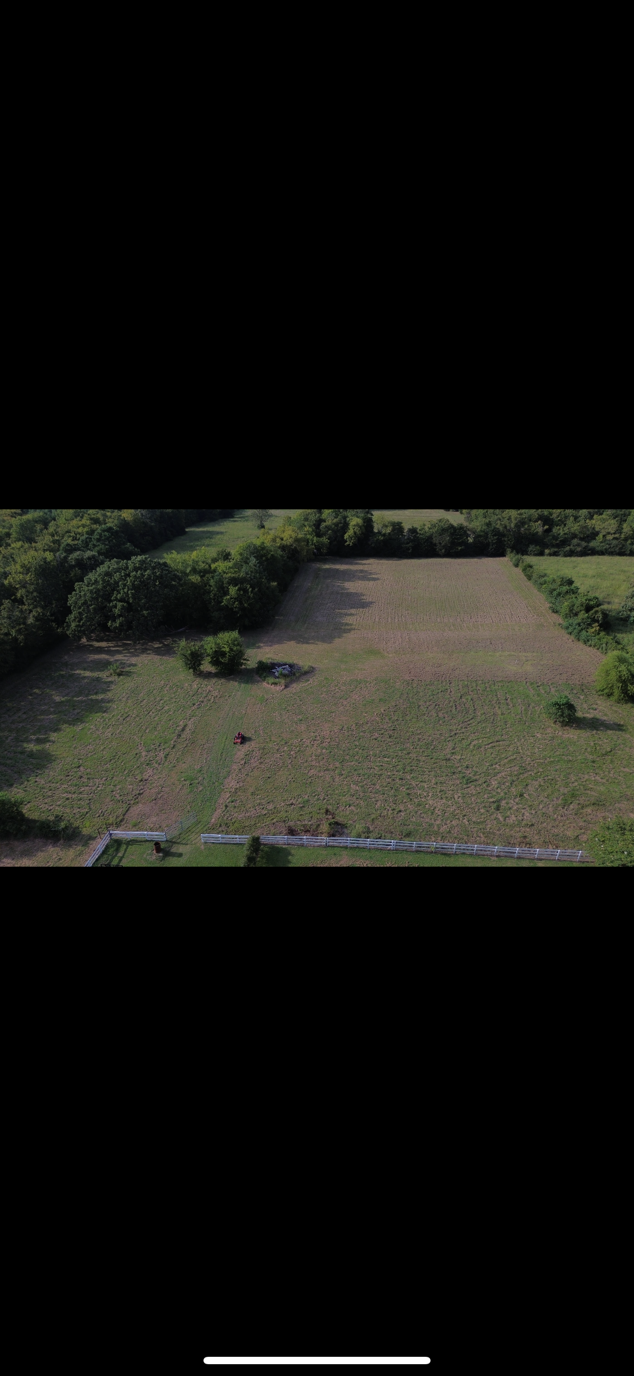 Aerial view of a grassy field bordered by trees, likely farmland, under a bright sky.