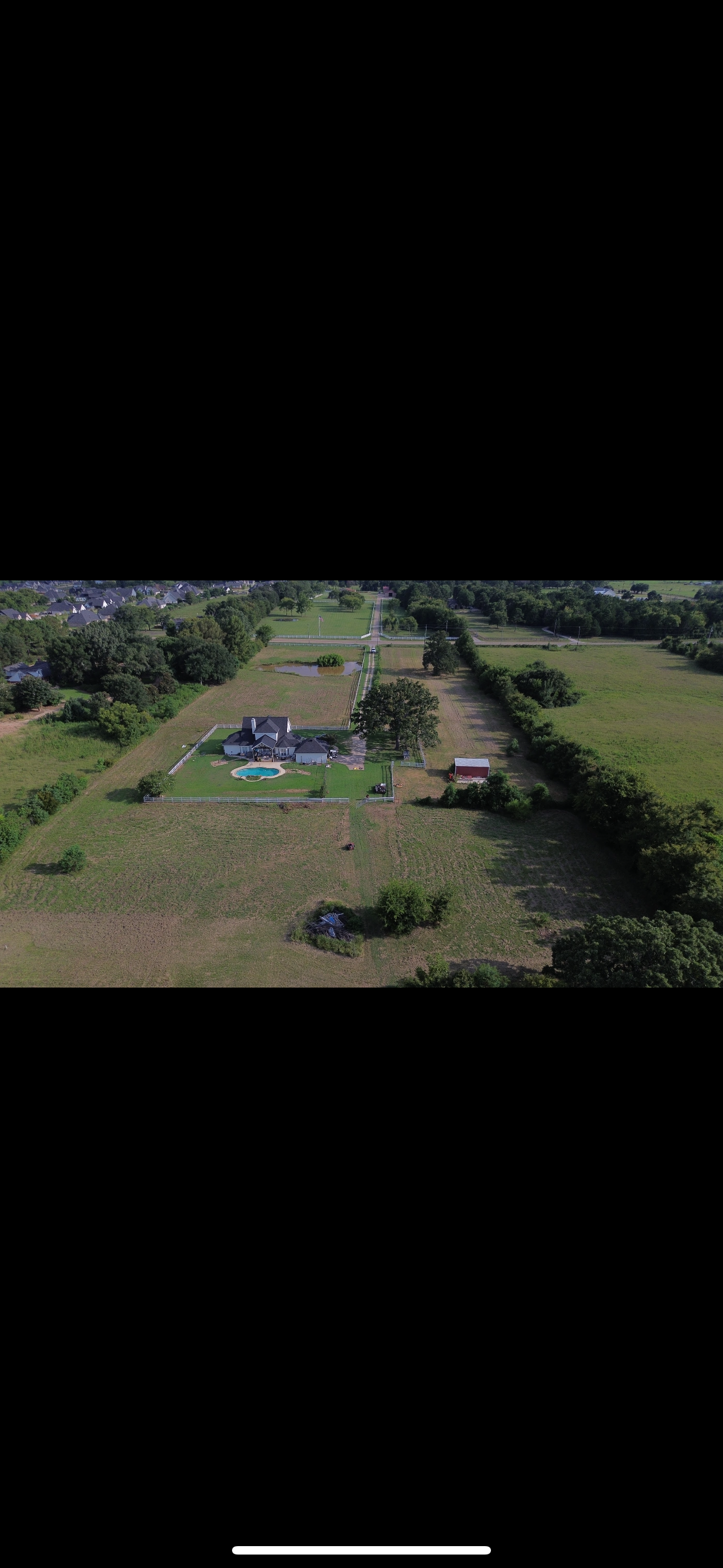 Aerial view of a house and surrounding property in a rural setting. Green grass and trees are visible.