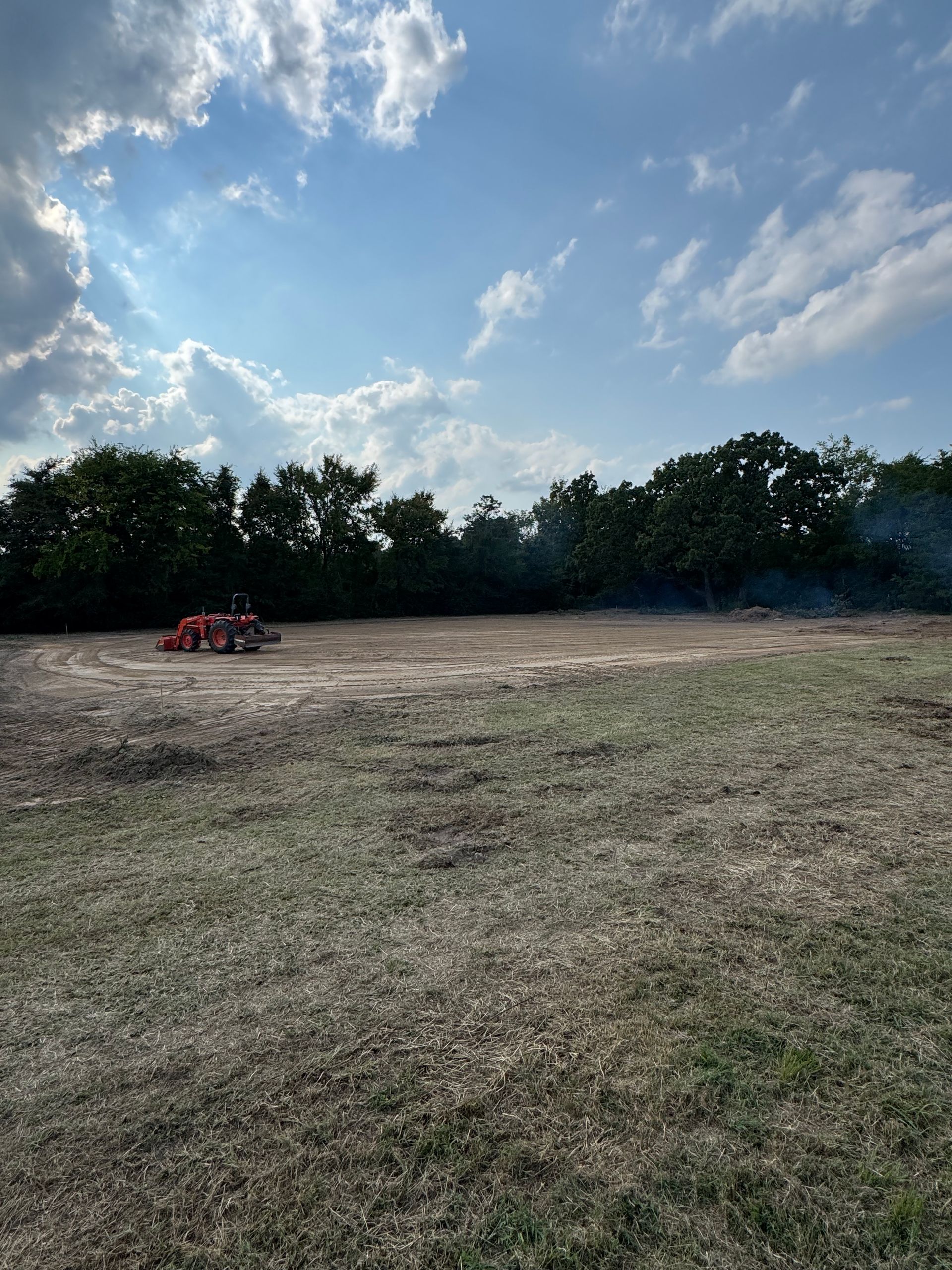 Field with a tractor and smoke rising in the distance under a partly cloudy sky.