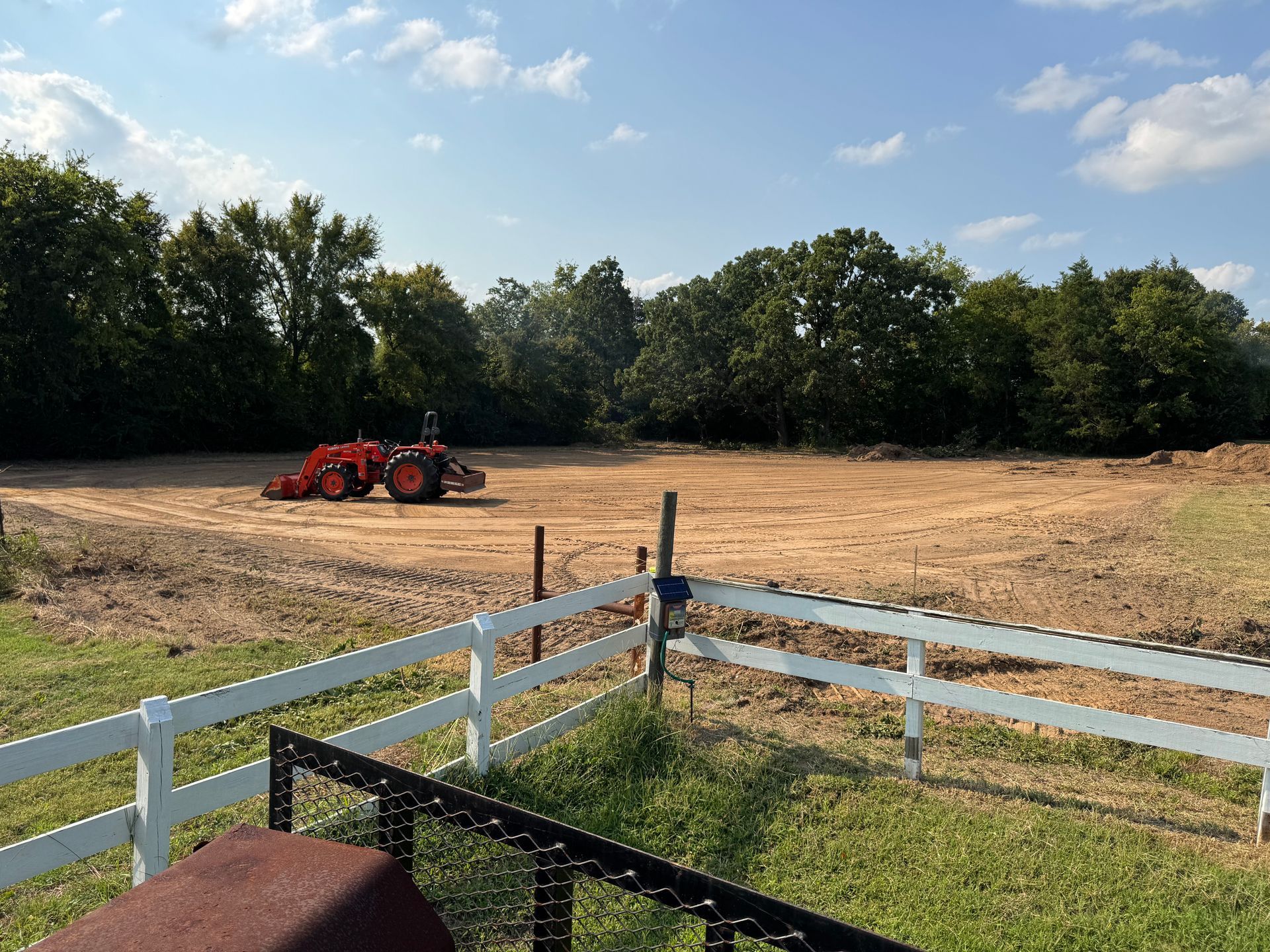 Tractor in a cleared field, white fence in foreground, trees in the distance, under a blue sky.
