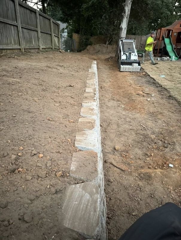 Retaining wall of stacked blocks in a dirt yard, with a construction worker and equipment in the background.