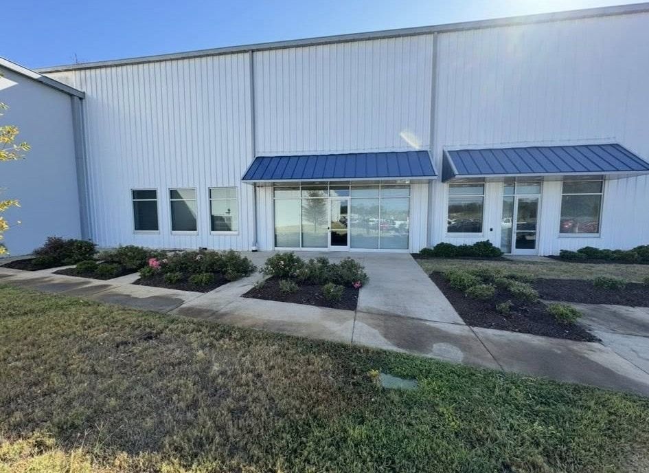 White industrial building entrance with blue awning, glass doors, and landscaping.