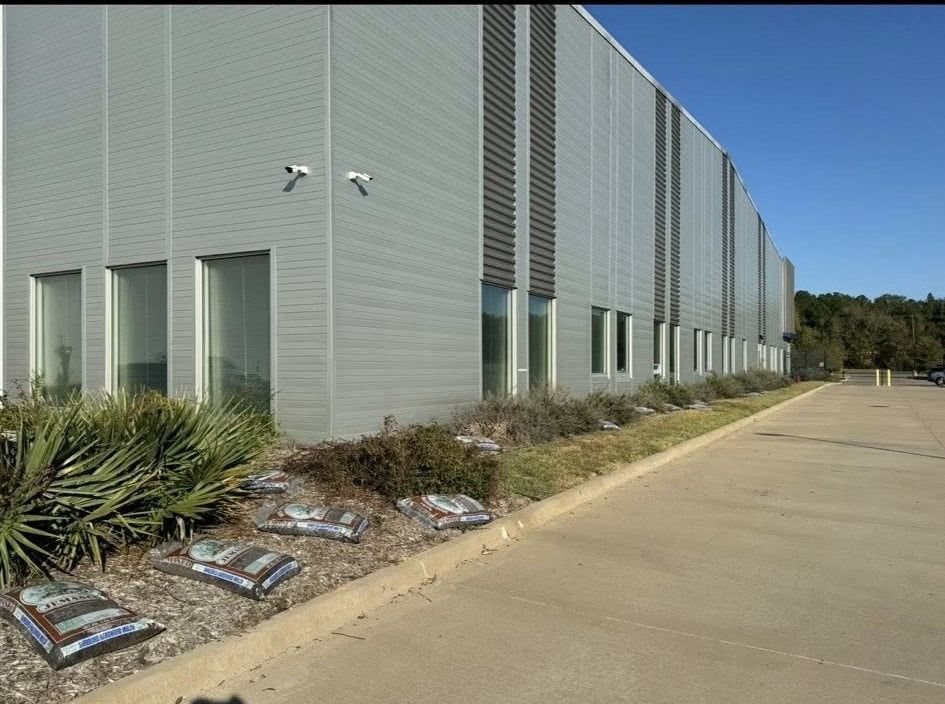 A modern, gray industrial building with a concrete parking area and landscaping under a blue sky.