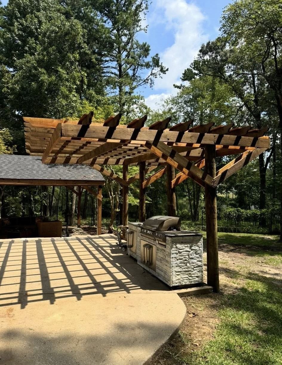 Outdoor kitchen with a pergola and grill, casting shadows on a concrete patio, surrounded by trees.