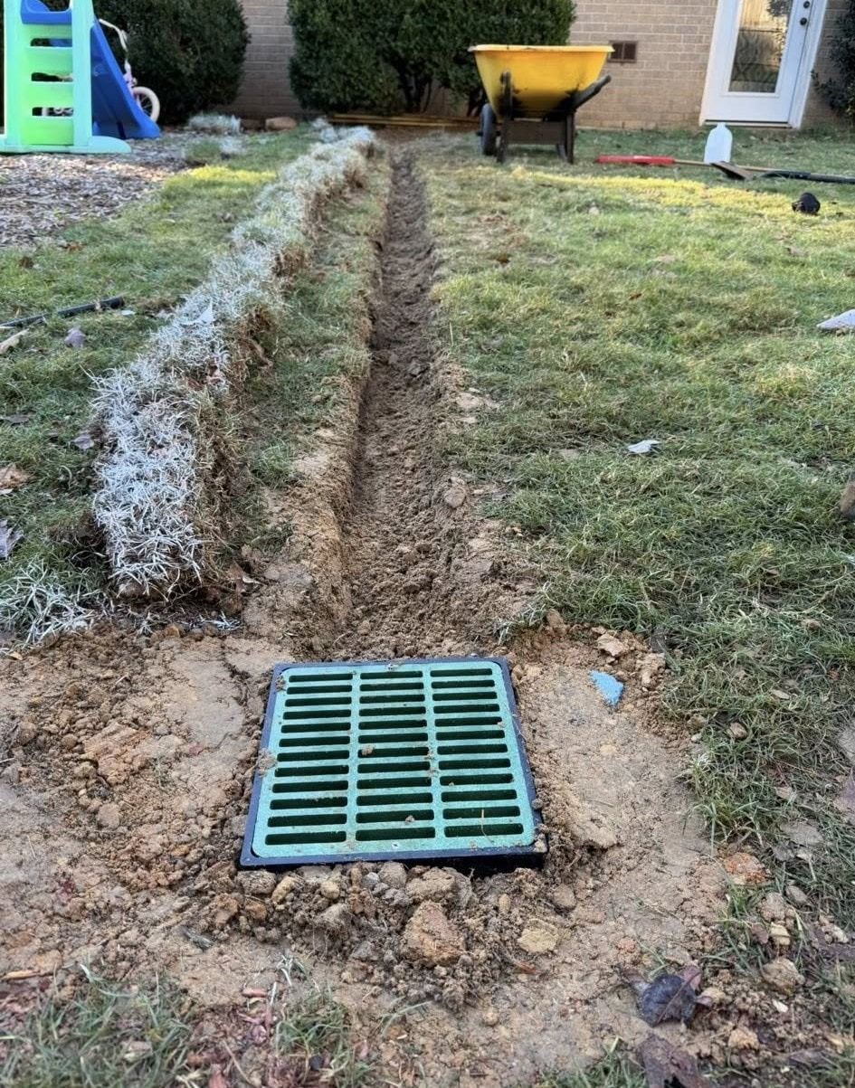 A green drain grate installed in a trench in a backyard, with a wheelbarrow and grass visible.