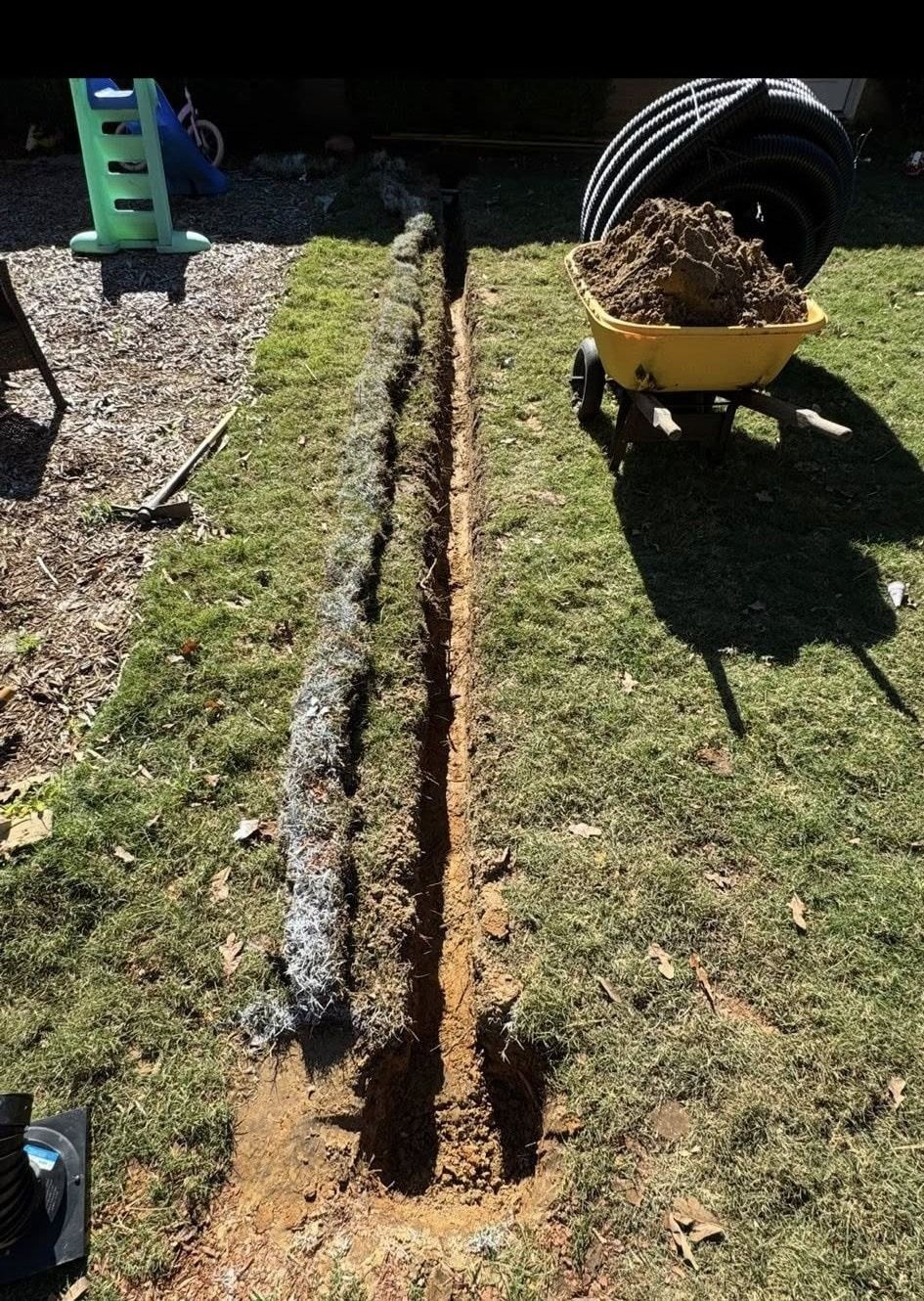 A narrow trench dug in grass, with wire mesh, a wheelbarrow of dirt, and conduit tubing visible.