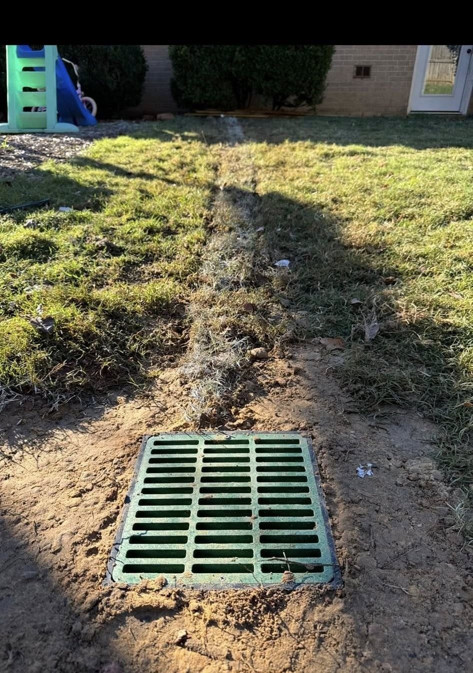 Green drain cover in dirt, with a trench leading to it in a grassy backyard.