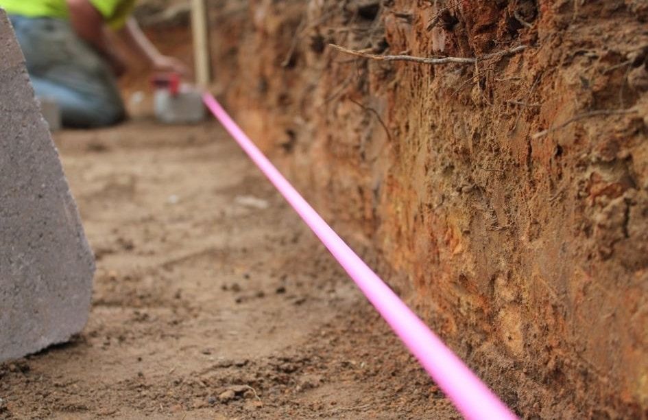 Pink utility line in trench, person kneeling in background, construction site.