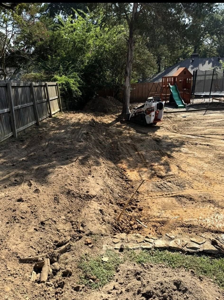A dirt backyard being excavated, a small tractor is visible near a tree, wooden fence and play structure are nearby.