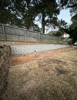 A retaining wall of gray blocks supports a backyard with dry grass, a wooden fence, and trees.