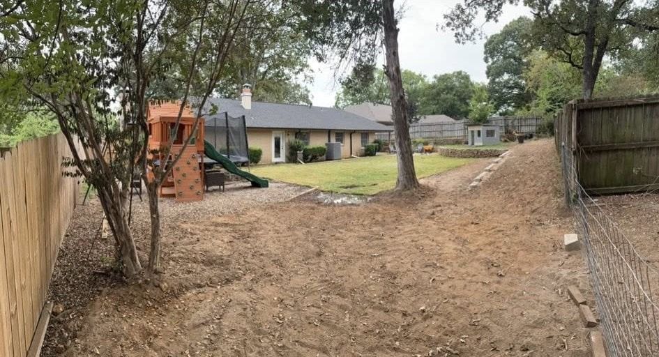 Backyard with a play structure, trampoline, and a house with a gray roof. Brown dirt covers much of the ground.