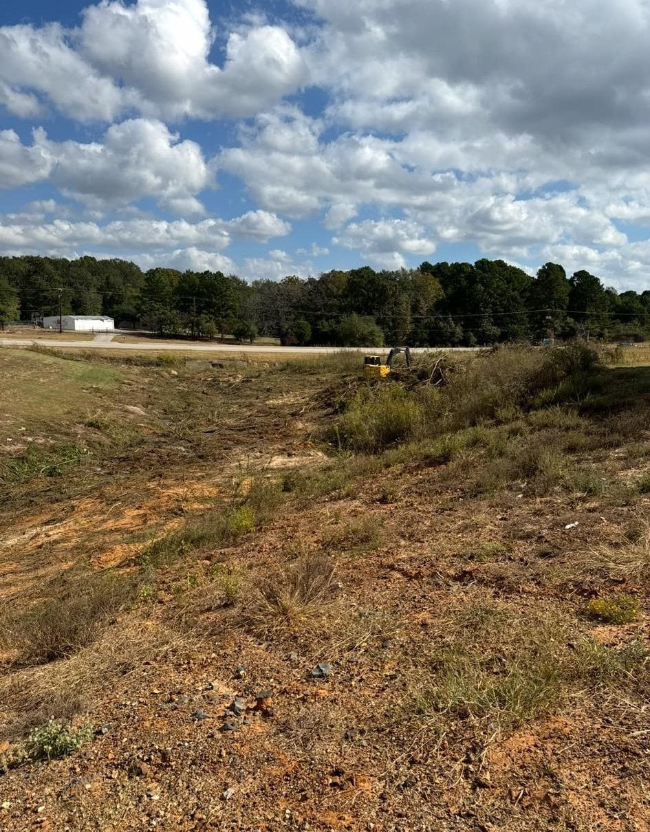 A dry, eroded ditch under a cloudy blue sky, with trees and a small vehicle in the distance.