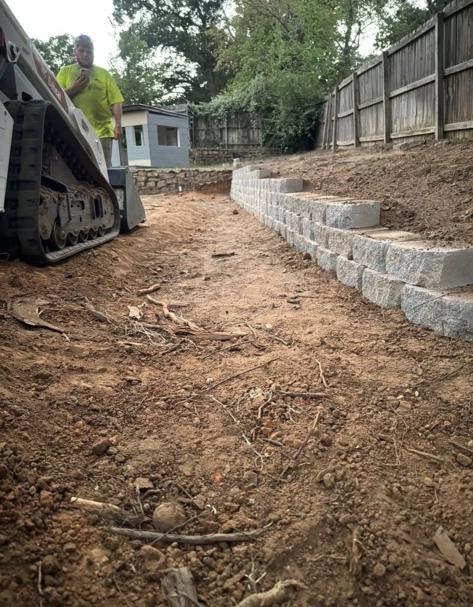 A construction site with a man operating a track loader near a retaining wall made of gray blocks.