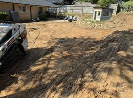 A construction site in a backyard with a mini excavator, dirt, and a building.