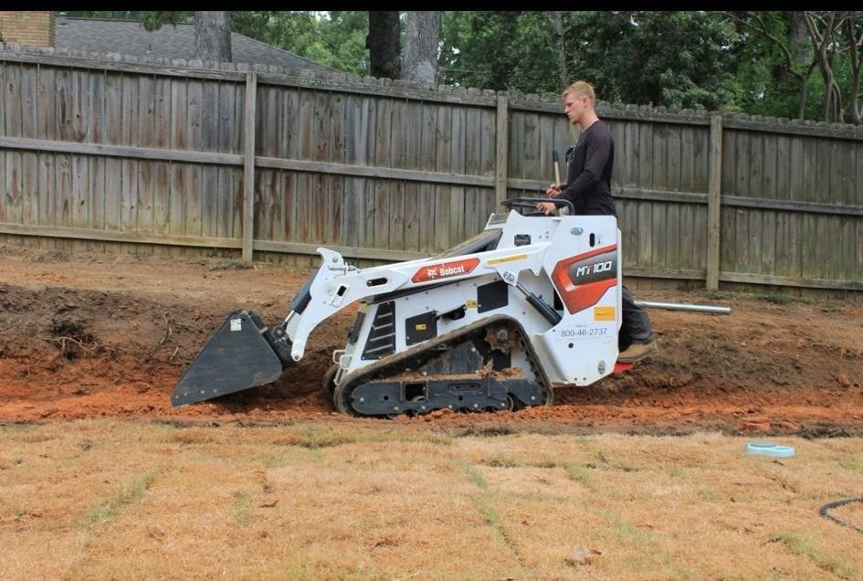 Man operating a white tracked skid steer on a hillside next to a wooden fence.