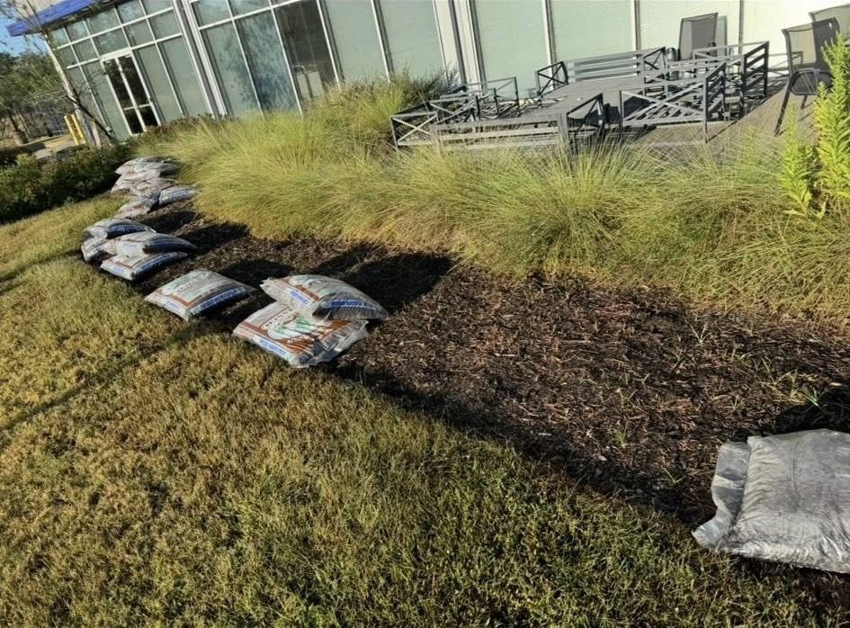 Bags of mulch in a row on a grassy hillside next to a building with tall grass and a seating area.