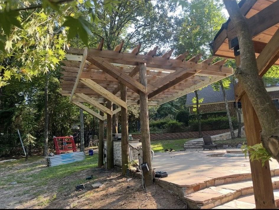 Pergola under construction, wood structure with angled supports, outdoors, trees, and stone patio.