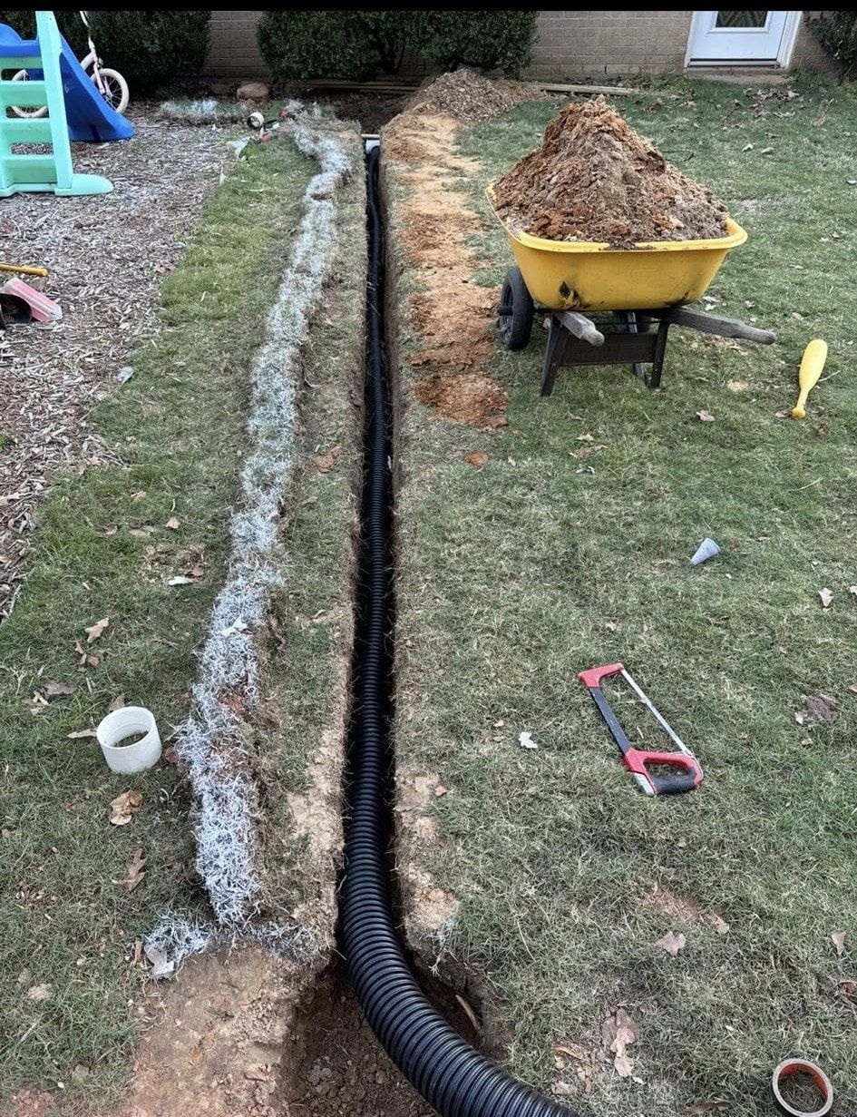 A black corrugated drain pipe in a trench, with a wheelbarrow of dirt, in a yard.