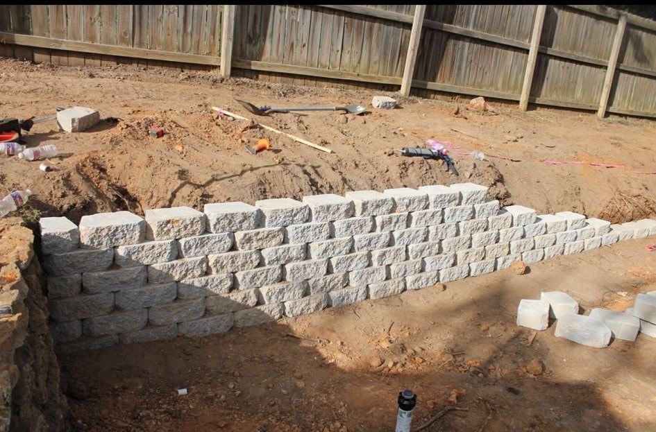 A partially built retaining wall made of gray stone blocks in a dirt yard, next to a wooden fence.