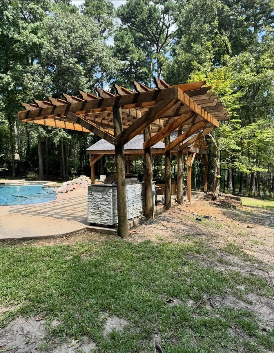 Wooden pergola over an outdoor kitchen near a pool, surrounded by trees and grass.