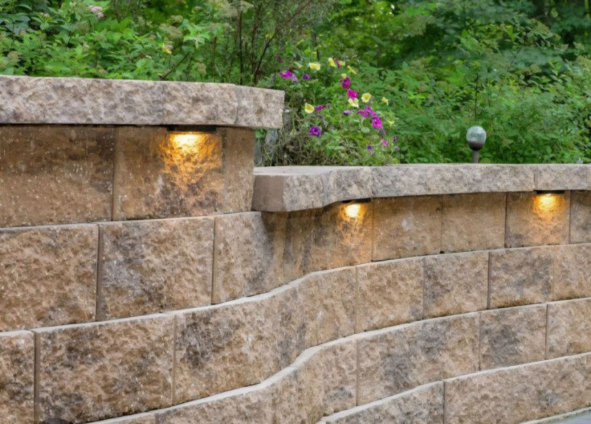 Stone retaining wall with built-in lights, curving down a hillside, with greenery in the background.