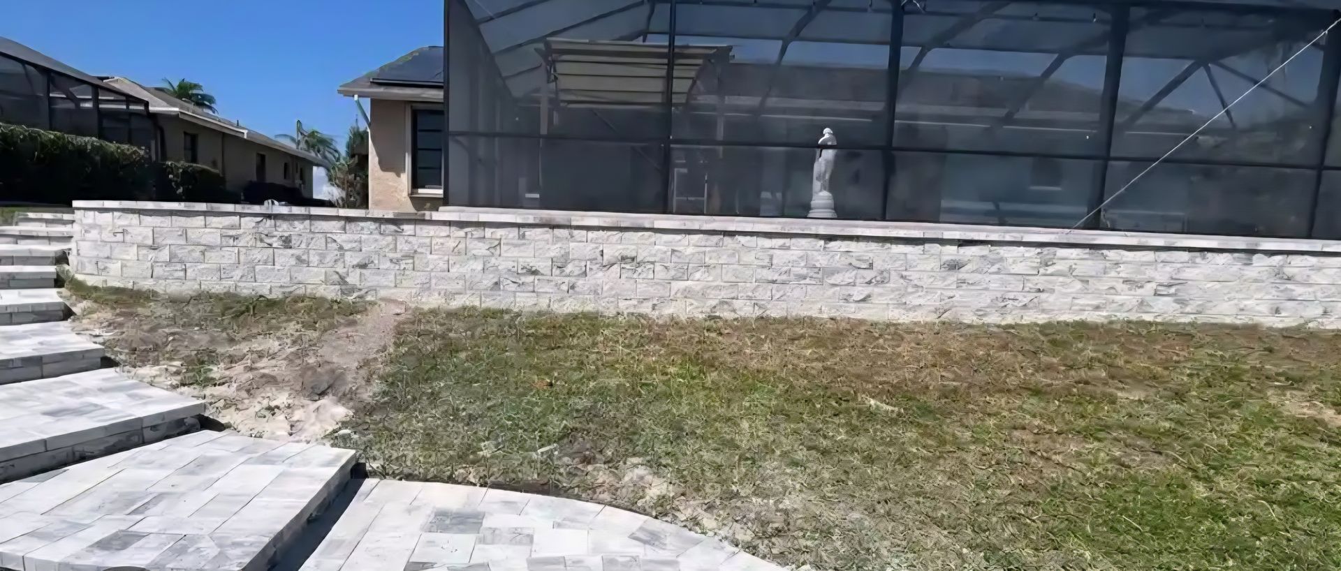 Stone wall with steps leading to a yard with patchy grass. A screened-in pool area is behind the wall.