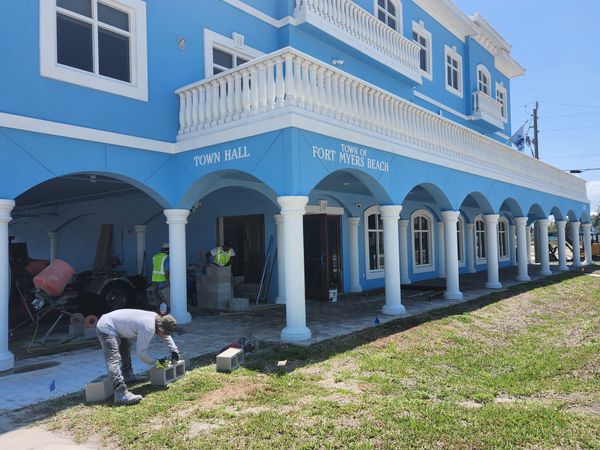 A man wearing a tool belt is pointing at something. Workers building in front of a bright blue building with white columns and trim.