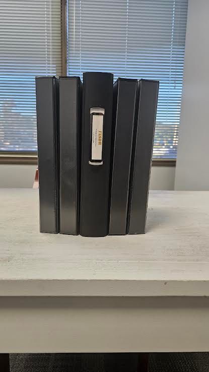 Black binders on a white table in front of a window with blinds. The center binder has a label.