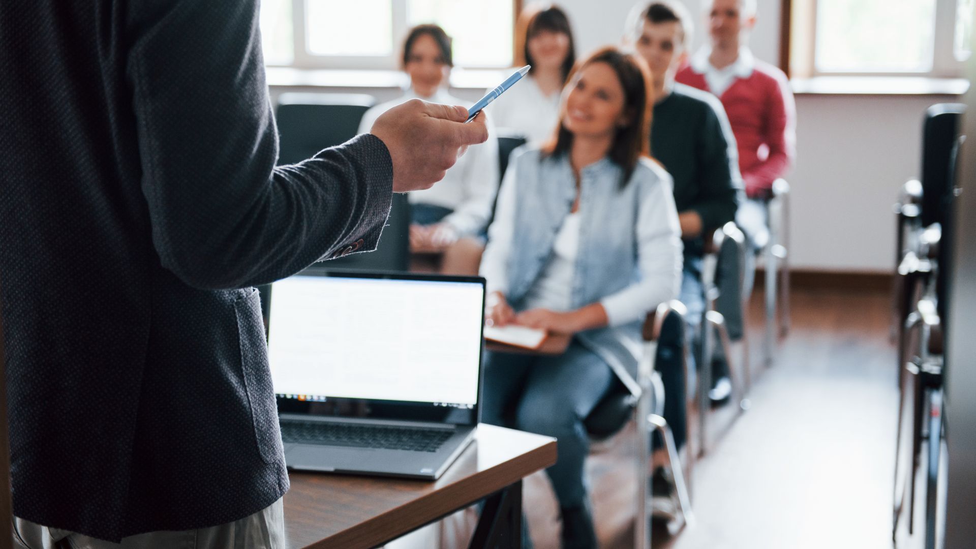 Person lecturing to a group in a classroom; laptop on table, students smiling.