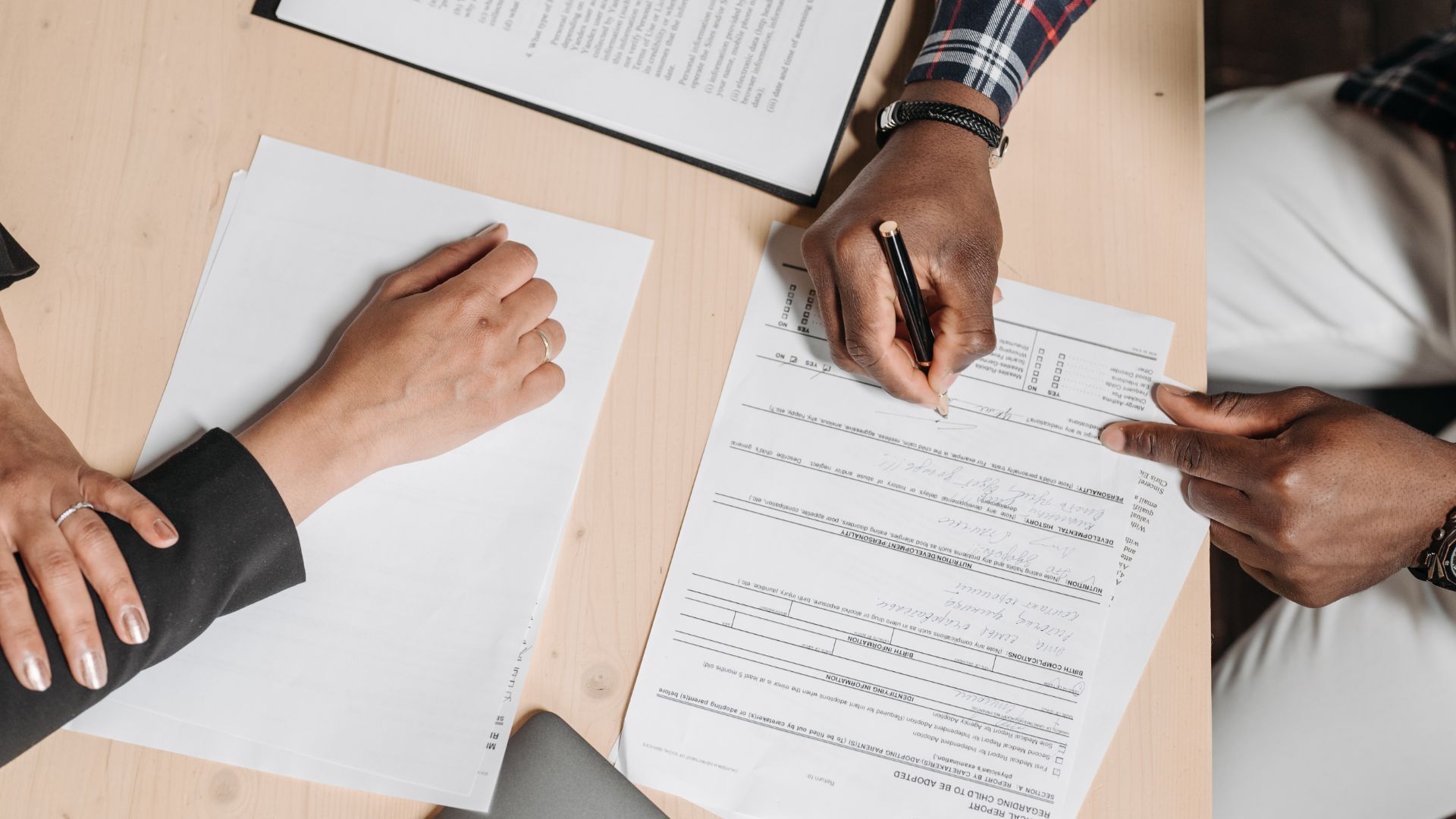 Hands signing a document, one hand pointing. Paperwork on a light wood table.