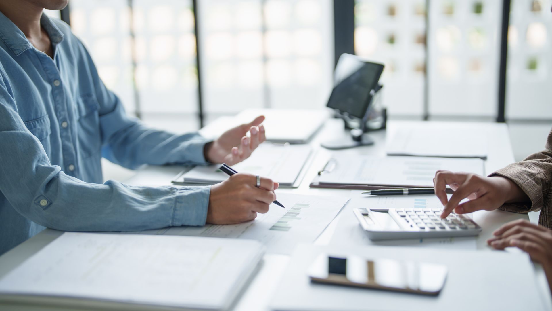 Two people at a desk review documents. One gestures, the other uses a calculator.