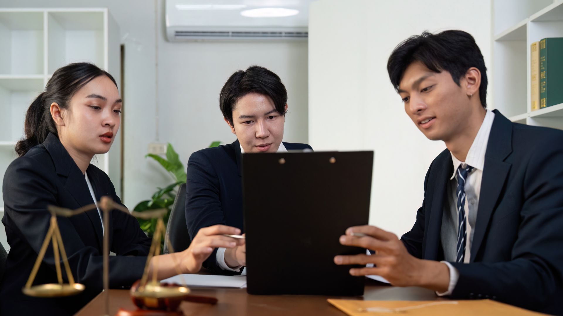 Three people in suits reviewing a document in an office, legal scale in the foreground.