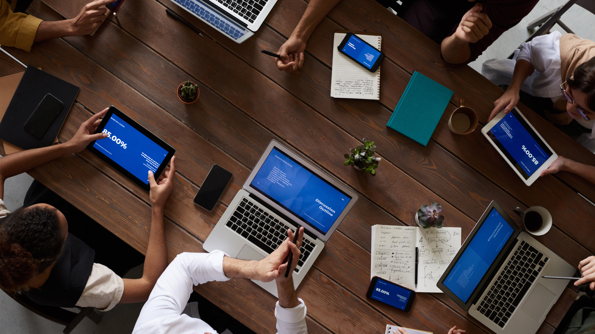 People at a wooden table with laptops, tablets, and phones, possibly a meeting.