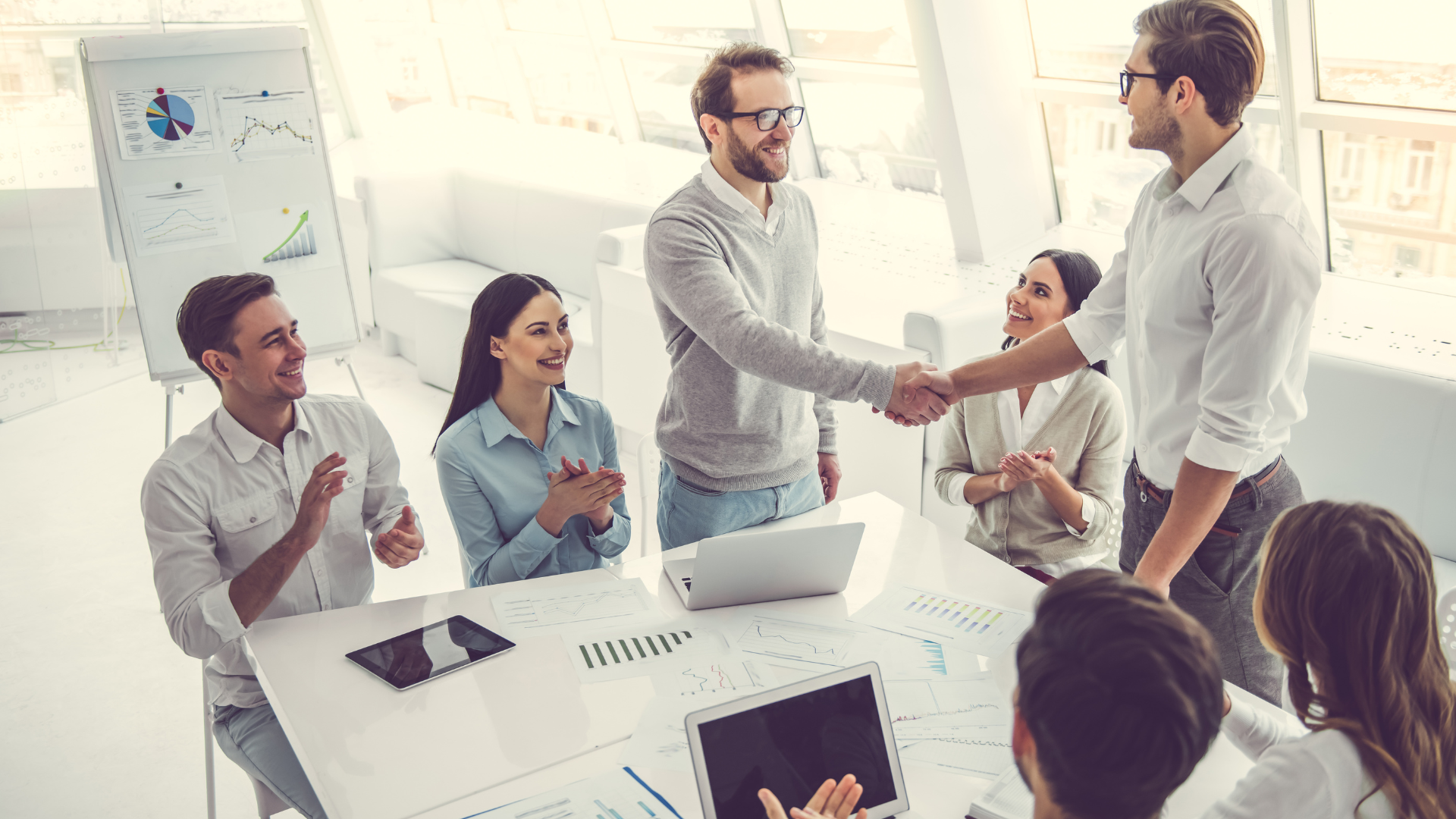 Business colleagues shake hands, others clap in a modern, bright office.