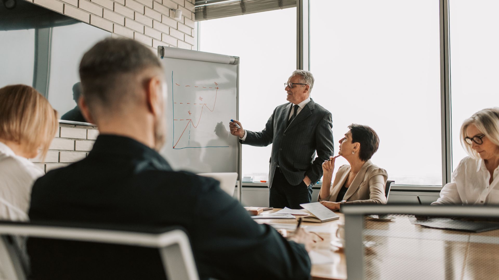 Business presentation: Man in suit points at whiteboard, facing a group in a conference room.