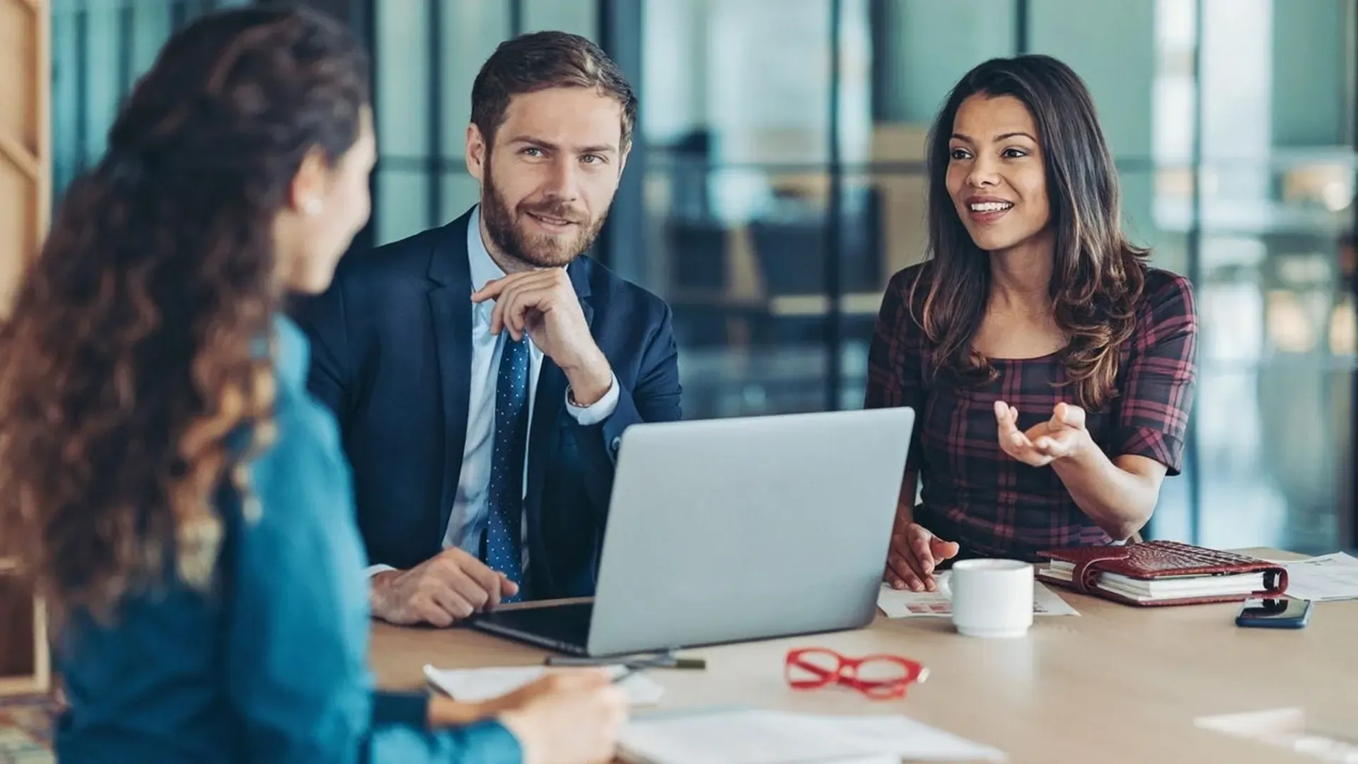 Three people at table; woman gesturing, man using laptop, and another woman taking notes.