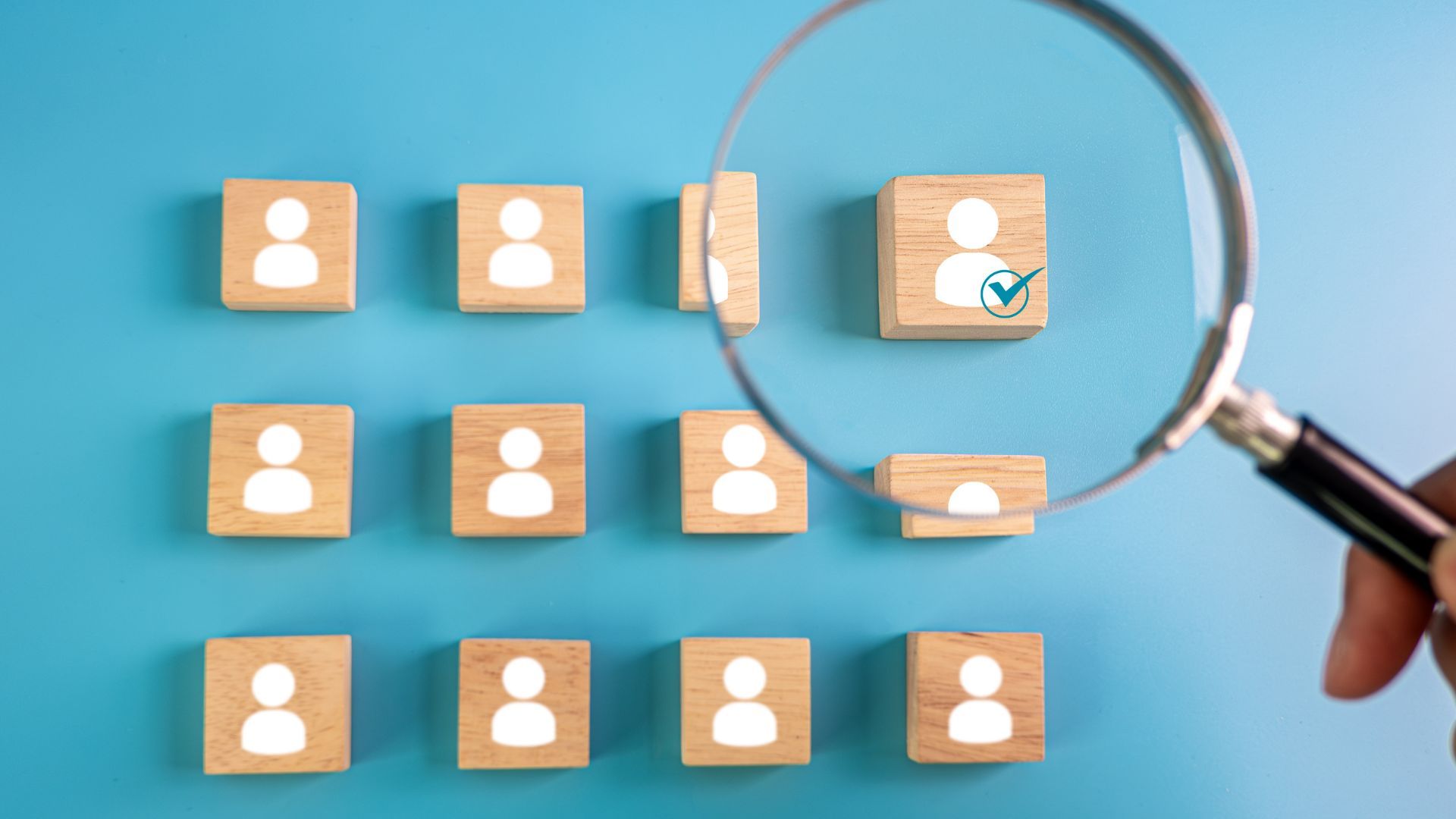 Magnifying glass over a wooden block with a person icon and a checkmark, on blue surface with other person icons.