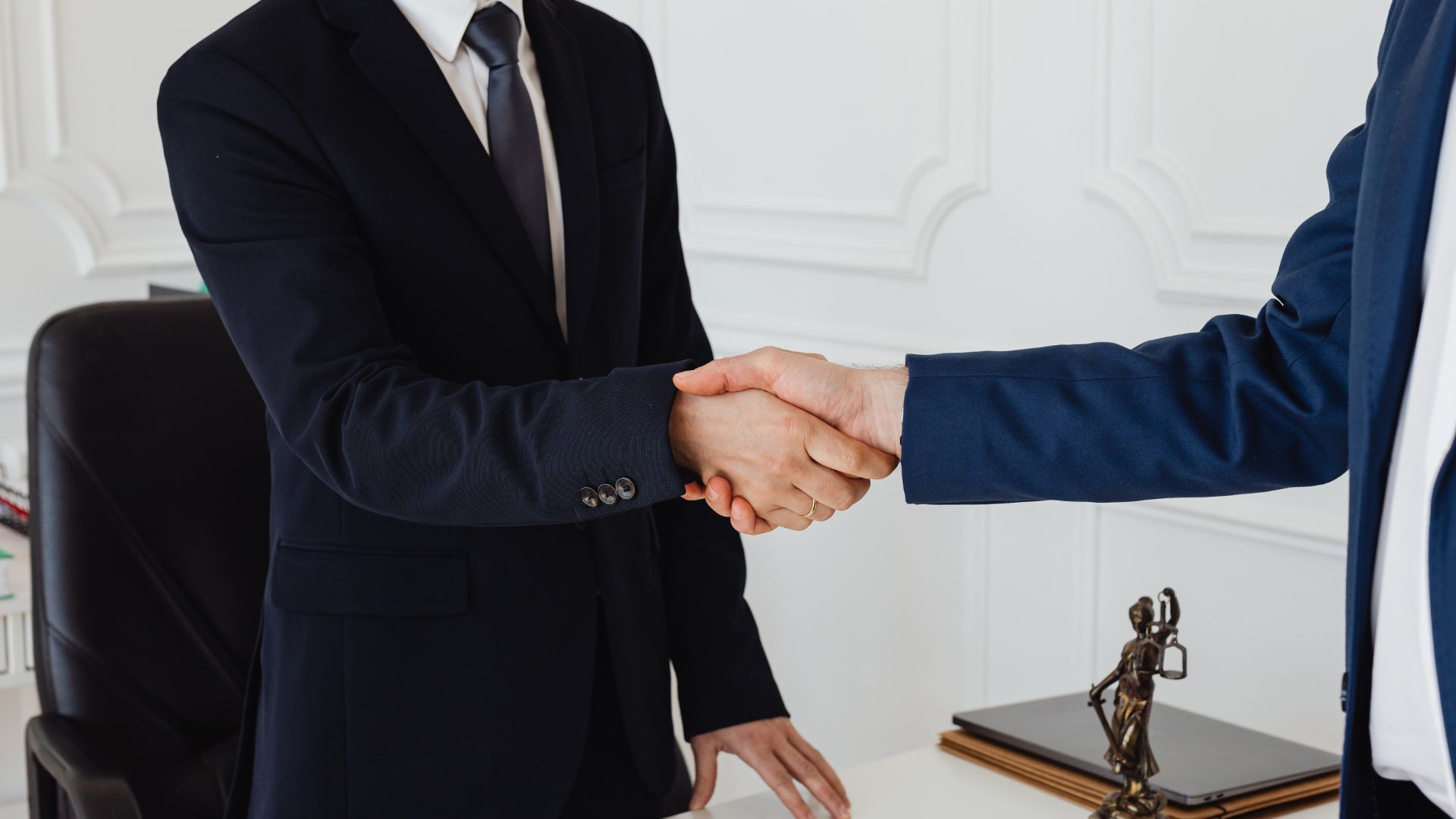 Two people in suits shaking hands in an office, symbolizing an agreement.