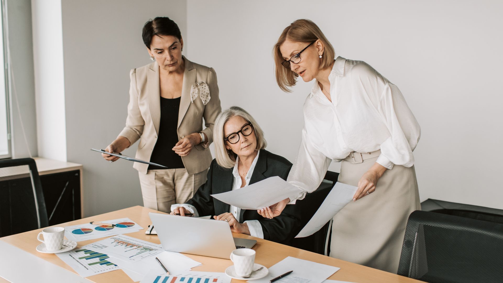 Three women in business attire reviewing documents and laptop at a desk in an office.