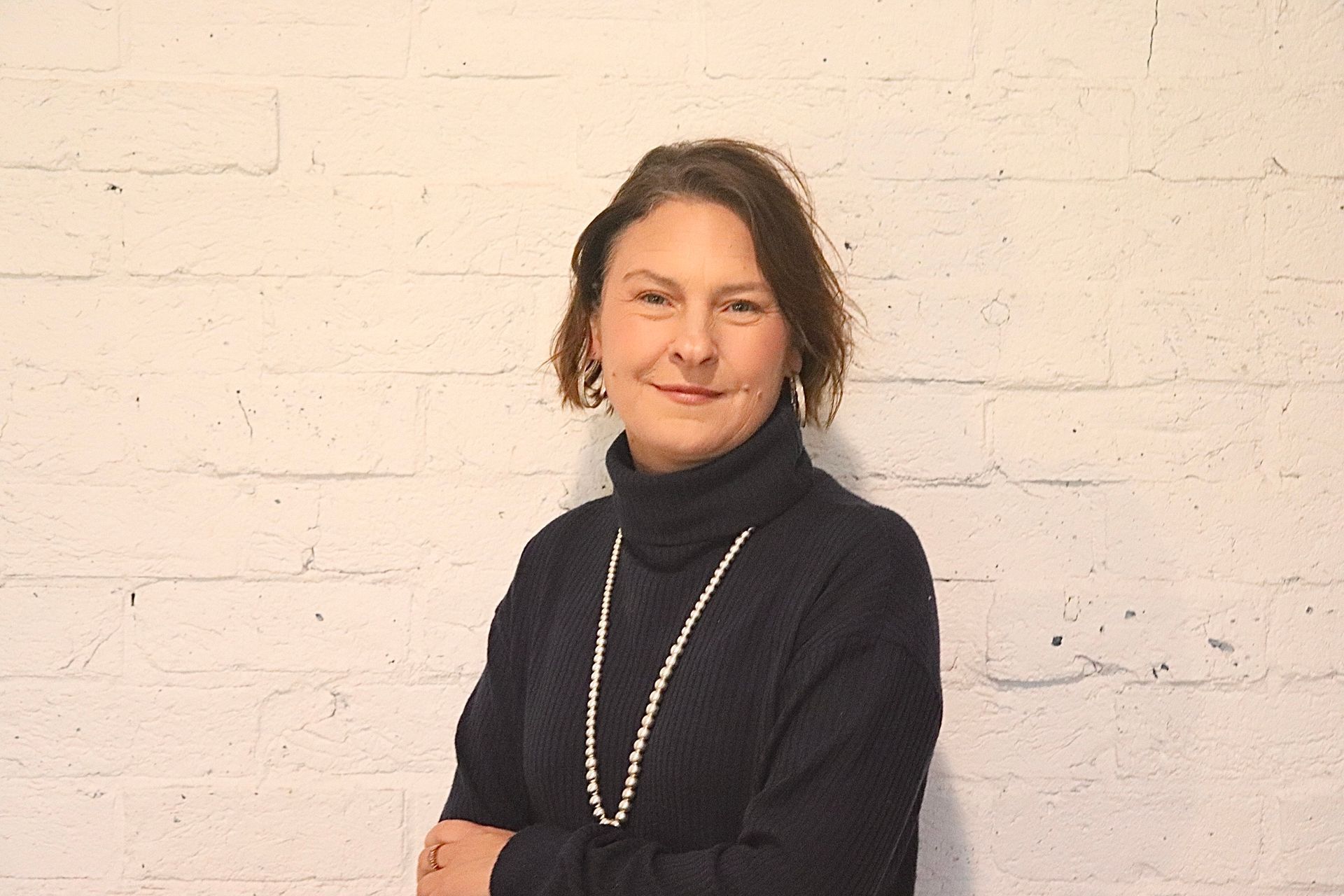 Tanya Leschev wearing a black turtleneck and pearls is standing in front of a white brick wall.