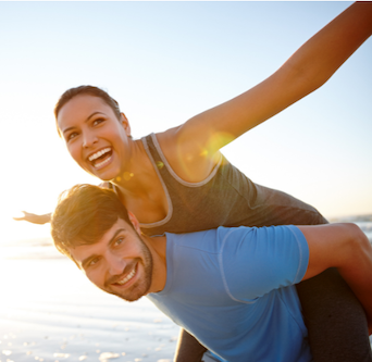 Man carrying a woman piggyback-style on a beach, both smiling. Sunlight.