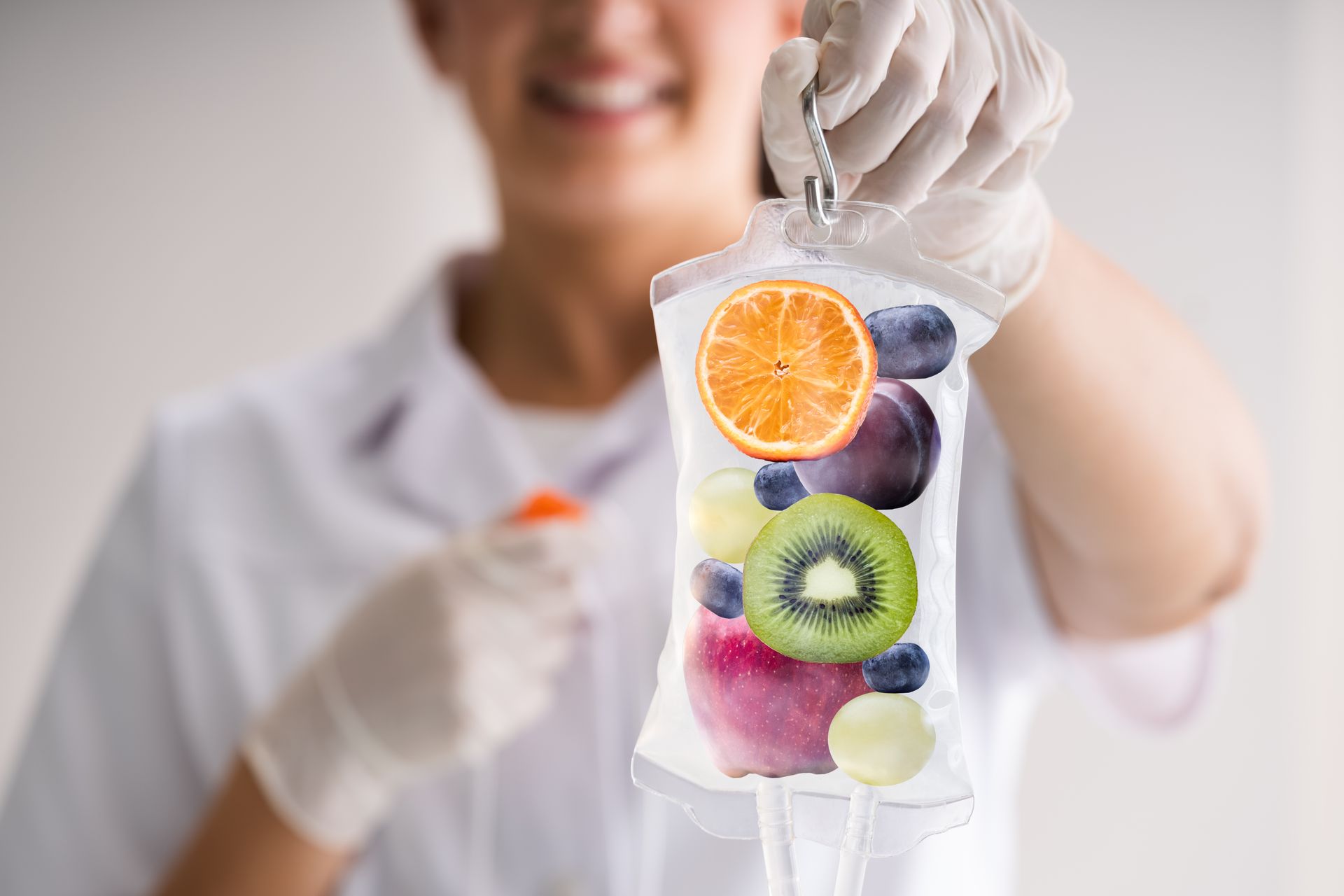 Photograph of a person wearing white gloves holding a clear IV bag filled with various fruits including an orange slice, kiwi, grapes, blueberries, and an apple. Photograph of a person wearing white gloves holding a clear IV bag filled with various fruits including an orange slice, kiwi, grapes, blueberries, and an apple.