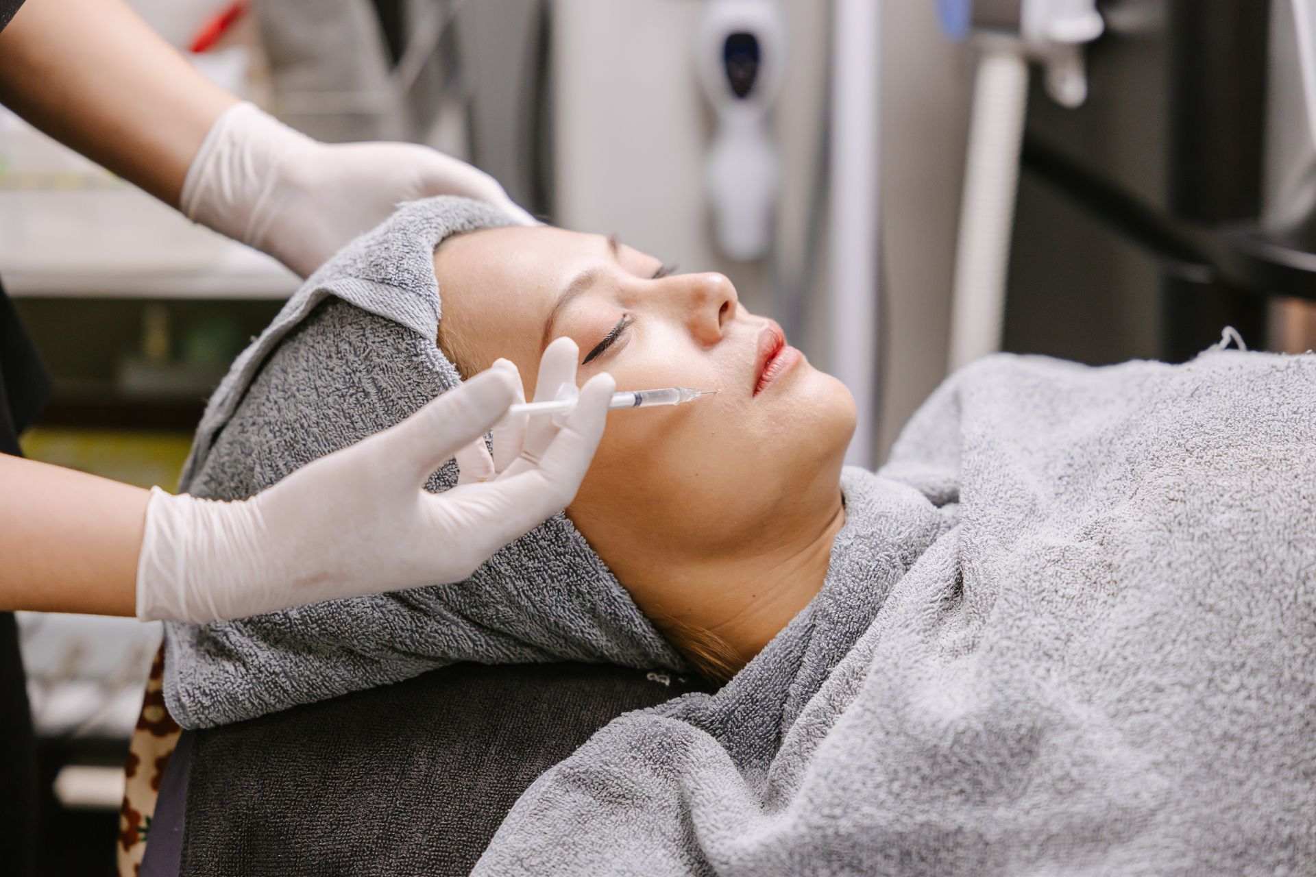 A person lying down, wrapped in a grey towel, receives a cosmetic injection in the cheek.
