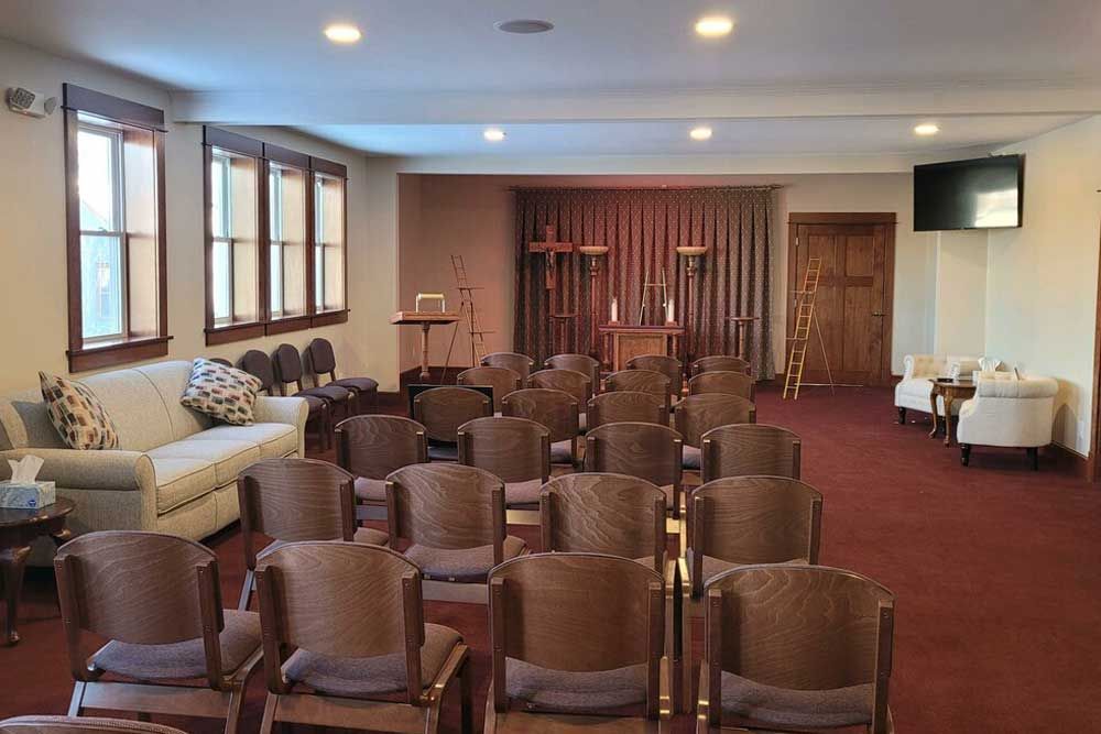 A quiet chapel room with rows of wooden chairs, a light-colored sofa, and a draped altar area in the background.
