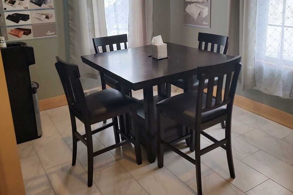 A dark-colored, square high-top dining table with four matching slatted-back chairs sits on a light-colored tiled floor.