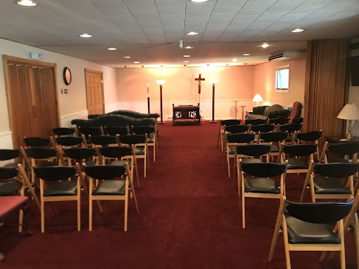 Interior of a chapel with rows of chairs facing a central altar featuring a cross on a red carpeted floor.