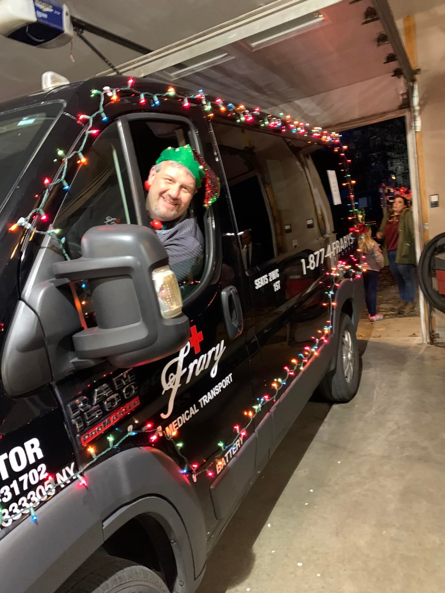 A person in a green elf hat smiles from the driver's seat of a black van decorated with colorful holiday lights.