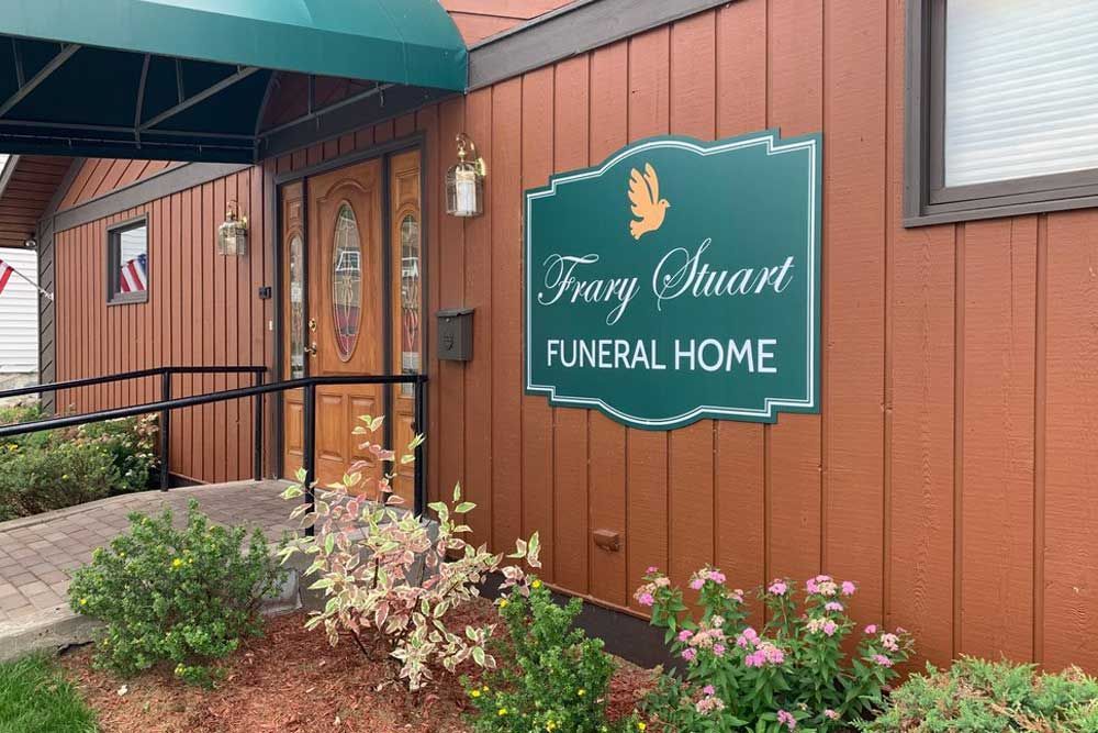 The exterior entrance of Frary Stuart Funeral Home, featuring a green sign with a dove, wood siding, and a ramp.