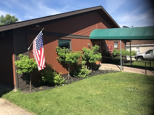 A brown building with a green awning, a US flag on the wall, and a patch of green grass in the foreground.